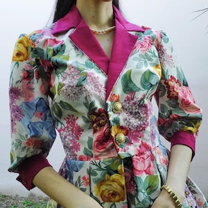 Puede incluir: Blazer con estampado floral sobre fondo blanco, con una vibrante mezcla de flores rosas, amarillas y azules. El blazer tiene cuello y puños fucsia, y está adornado con botones dorados. Las mangas son de tres cuartos.