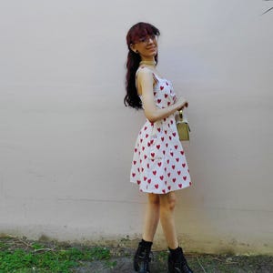 May include: A white sundress with a pattern of red hearts. The dress has a fitted bodice and a flared skirt. The person is wearing black boots and holding a small green purse. The background is a light pink wall and green grass.