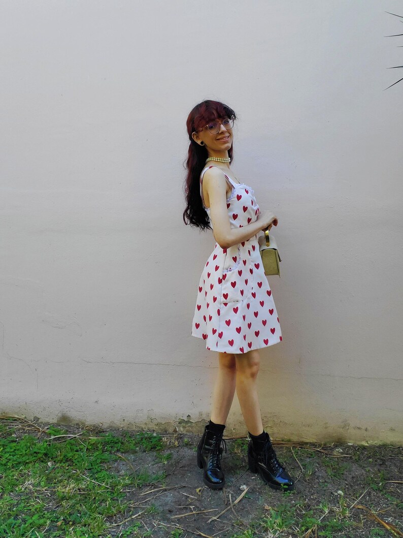 May include: A white sundress with a pattern of red hearts. The dress has a fitted bodice and a flared skirt. The person is wearing black boots and holding a small green purse. The background is a light pink wall and green grass.