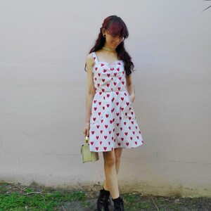 May include: A white two-piece outfit with a red heart pattern. The top has a square neckline and the skirt is a mini-skirt. The outfit is paired with black boots and a small, light green handbag. The background is a light pink wall and green grass.