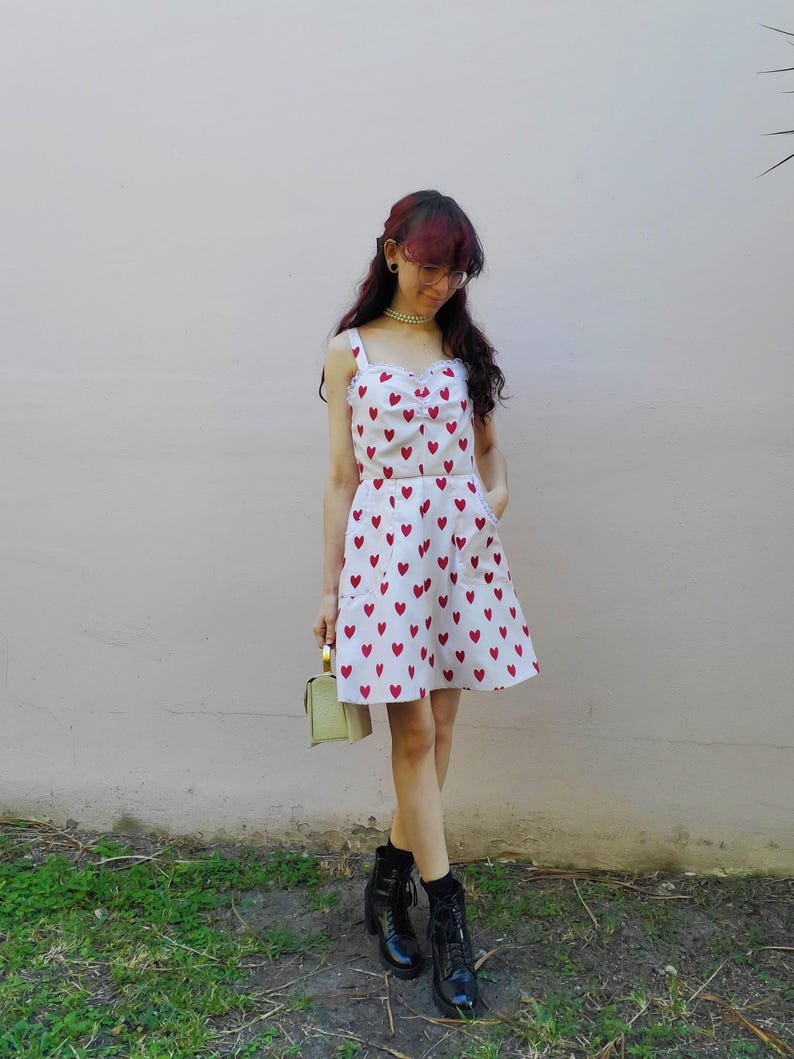 May include: A white two-piece outfit with a red heart pattern. The top has a square neckline and the skirt is a mini-skirt. The outfit is paired with black boots and a small, light green handbag. The background is a light pink wall and green grass.