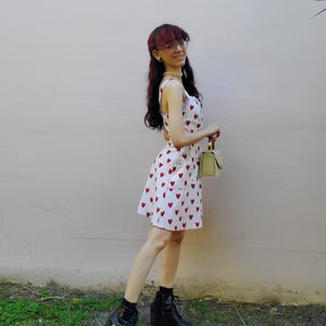May include: White sundress with red heart pattern. The dress has a scoop neckline and a fitted silhouette. The person is wearing black ankle boots with a chunky heel and a small, light green handbag.