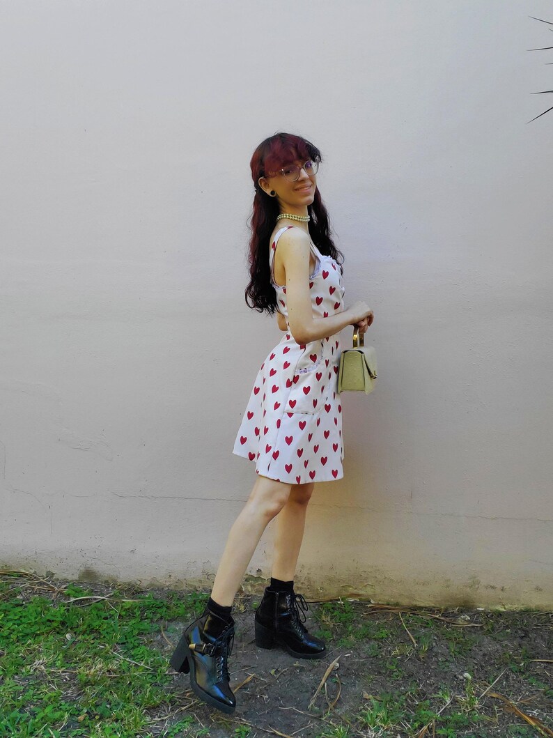 May include: White sundress with red heart pattern. The dress has a scoop neckline and a fitted silhouette. The person is wearing black ankle boots with a chunky heel and a small, light green handbag.