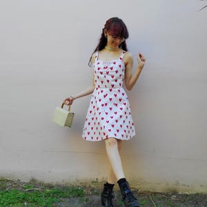 May include: A white sundress with red heart patterns. The dress has a sweetheart neckline and spaghetti straps. The person is wearing black boots and holding a light green handbag. The background is a light pink wall.