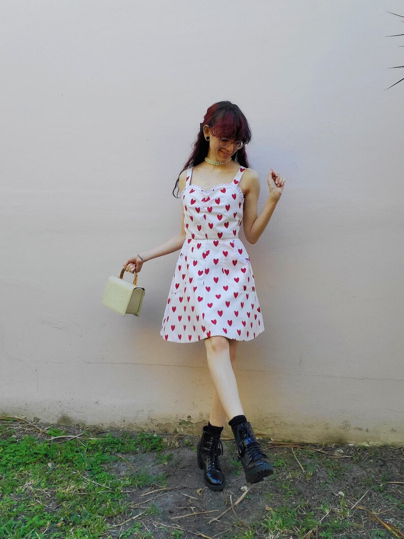 May include: A white sundress with red heart patterns. The dress has a sweetheart neckline and spaghetti straps. The person is wearing black boots and holding a light green handbag. The background is a light pink wall.