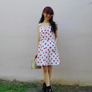 May include: A white dress with red heart patterns. The dress has a fitted bodice with a sweetheart neckline and spaghetti straps. The skirt is a-line with pockets. The person is wearing black boots and holding a light green purse.