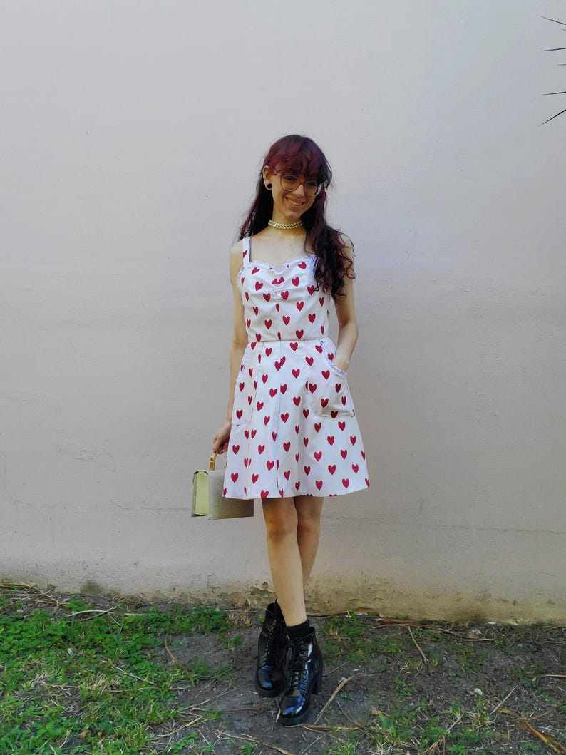 May include: A white dress with red heart patterns. The dress has a fitted bodice with a sweetheart neckline and spaghetti straps. The skirt is a-line with pockets. The person is wearing black boots and holding a light green purse.
