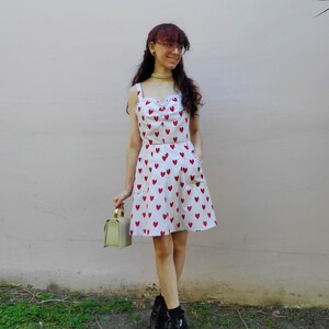 May include: A two-piece outfit featuring a white top and skirt, both adorned with a pattern of red hearts. The top has a sweetheart neckline and the skirt has pockets. The outfit is paired with black boots and a small green handbag.