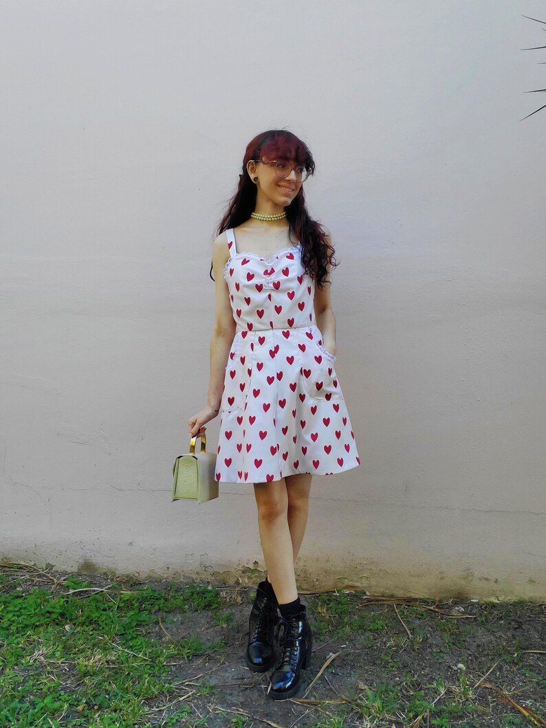 May include: A two-piece outfit featuring a white top and skirt, both adorned with a pattern of red hearts. The top has a sweetheart neckline and the skirt has pockets. The outfit is paired with black boots and a small green handbag.
