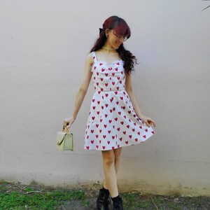 May include: A white dress with red heart patterns. The dress has a sweetheart neckline and spaghetti straps. The person is wearing black boots and holding a small, light green handbag.