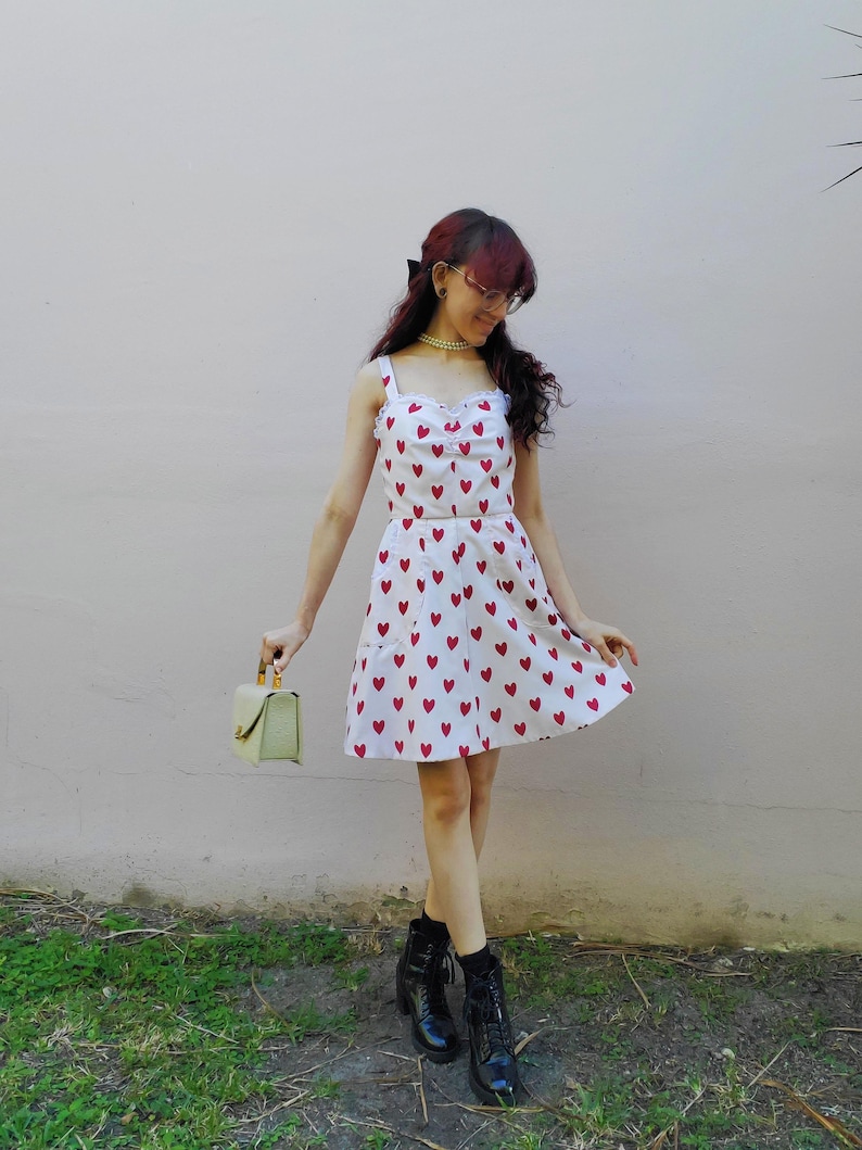 May include: A white dress with red heart patterns. The dress has a sweetheart neckline and spaghetti straps. The person is wearing black boots and holding a small, light green handbag.