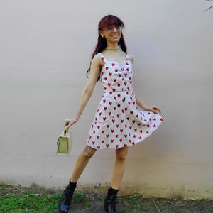 May include: A white sundress with a red heart pattern. The dress has a sweetheart neckline and spaghetti straps. The person is wearing black boots and holding a light green handbag.