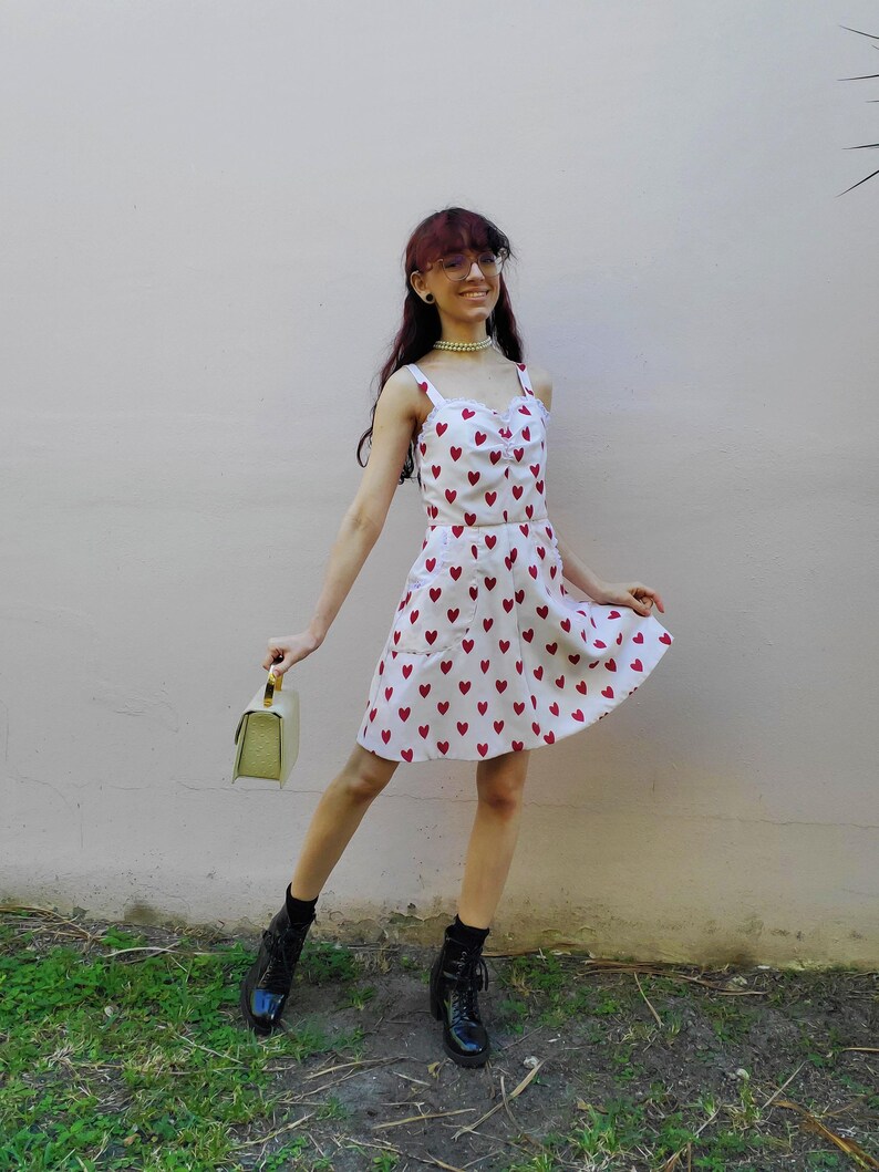 May include: A white sundress with a red heart pattern. The dress has a sweetheart neckline and spaghetti straps. The person is wearing black boots and holding a light green handbag.