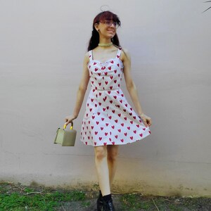 May include: A white sundress with red heart patterns. The dress has a scoop neckline, spaghetti straps, and a flared skirt. The person is wearing black boots and holding a small, olive-green handbag with a gold handle.