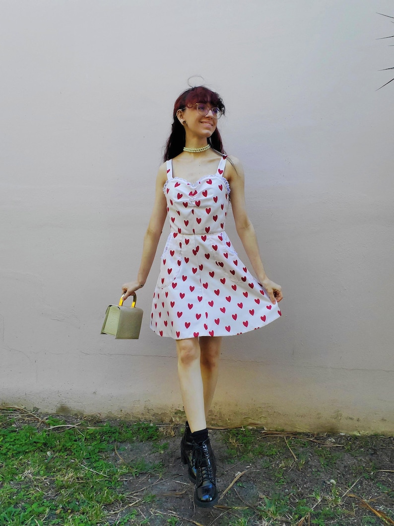 May include: A white sundress with red heart patterns. The dress has a scoop neckline, spaghetti straps, and a flared skirt. The person is wearing black boots and holding a small, olive-green handbag with a gold handle.