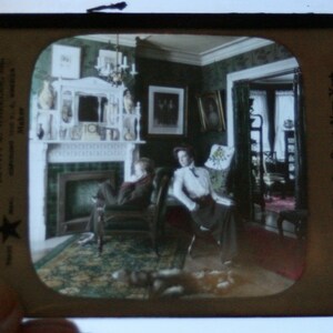 May include: A vintage stereoscopic image of a living room interior with two people sitting in chairs. The room features a fireplace, a large window, and a patterned rug. The text "120 and 122 West 31st Street, New York" is visible in the lower right corner.