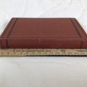 May include: A vintage, rectangular book with a brown cover and black stripes along the edges. The book is placed next to a wooden ruler. The text on the book's spine is partially visible. The ruler measures approximately 7 inches.
