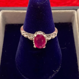 May include: A silver ring with a large oval ruby gemstone. The ring is set on a blue velvet display inside a jewelry box. The band features a decorative filigree design on either side of the ruby.