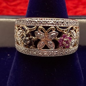 May include: A gold-toned ring with a floral design. The ring features a band of small diamonds and intricate cut-out patterns. The design includes flower and butterfly shapes, with accents of black, pink, and clear gemstones. The ring is set against a dark blue background.