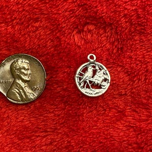 May include: A silver-colored charm with a bird design, next to a 1965 penny on a red background. The charm features two birds perched on a branch within a circular frame. The penny shows a profile of a man.