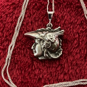 May include: Silver necklace with a pendant featuring a detailed profile of a woman wearing a winged hat with floral accents.