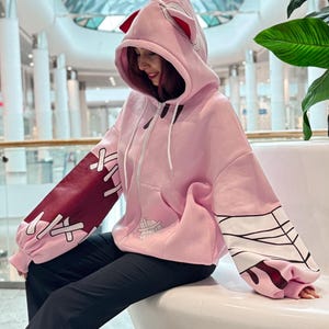 May include: Pink zip-up hoodie with a hood featuring red accents and ears. The sleeves have burgundy and white designs. The hoodie has a front pocket and white drawstrings. The person is wearing black pants.