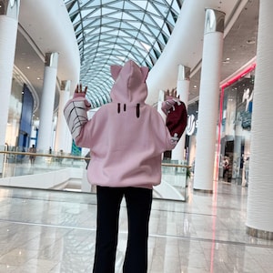 May include: A light pink hoodie with cat ears on the hood. The sleeves have a white spiderweb design and a burgundy color. The person is wearing black pants. The background is a modern shopping mall.