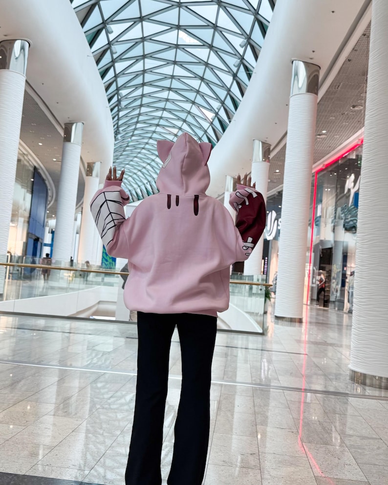 May include: A light pink hoodie with cat ears on the hood. The sleeves have a white spiderweb design and a burgundy color. The person is wearing black pants. The background is a modern shopping mall.