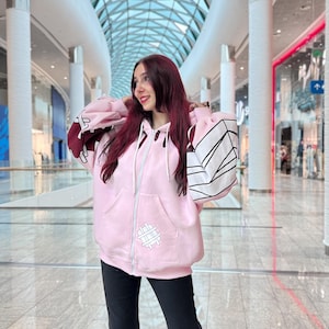 May include: A light pink zip-up hoodie with white and black geometric designs on the sleeves and a small white graphic on the front. The hoodie has a hood and two front pockets. The person is wearing black pants.