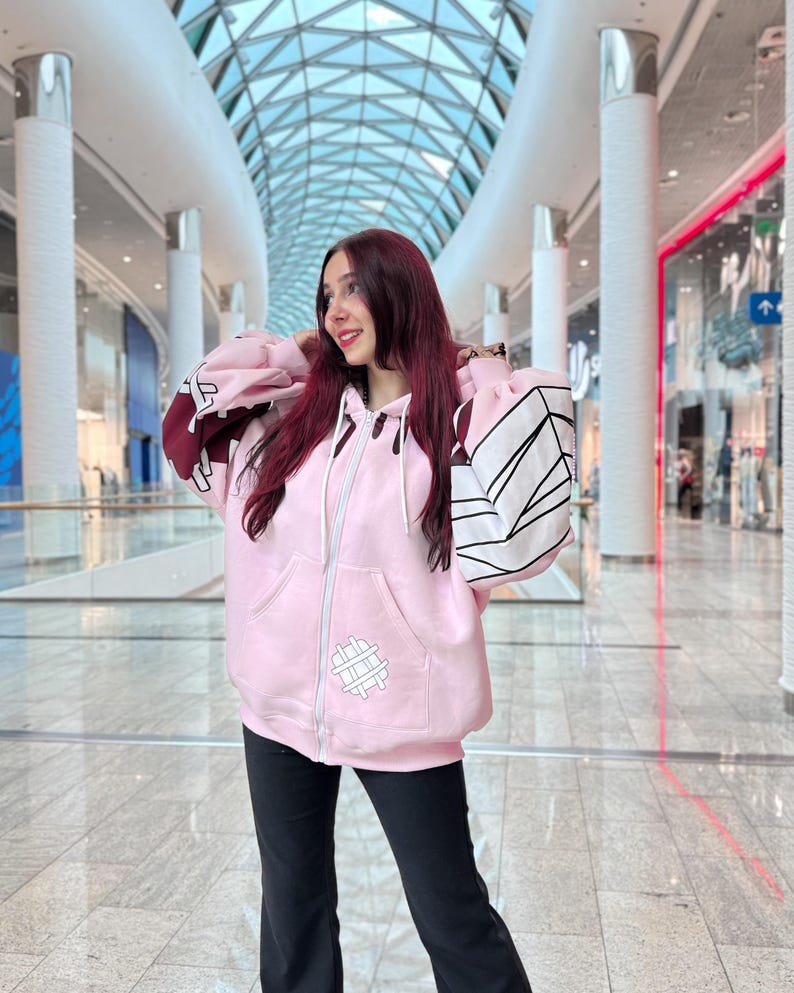 May include: A light pink zip-up hoodie with white and black geometric designs on the sleeves and a small white graphic on the front. The hoodie has a hood and two front pockets. The person is wearing black pants.