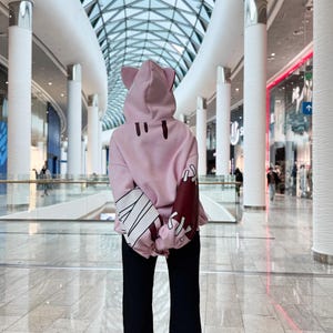 May include: A light pink hoodie with cat ears on the hood and black pants. The hoodie has dark red and white accents on the sleeves. The person is standing in a modern shopping mall with a glass ceiling.