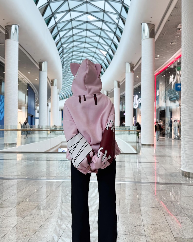 May include: A light pink hoodie with cat ears on the hood and black pants. The hoodie has dark red and white accents on the sleeves. The person is standing in a modern shopping mall with a glass ceiling.