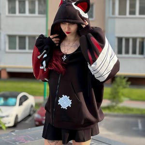 Op de afbeelding: Zwarte hoodie met kattenoordesign. De hoodie heeft rode accenten op de mouwen en capuchon, met witte strepen en een opgestikte zak. De rits is zichtbaar aan de voorkant. De mouwen zijn lang en oversized.