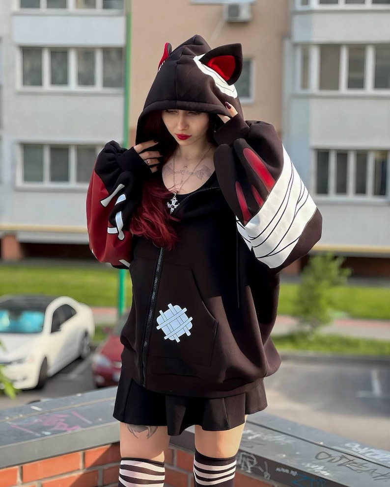 Op de afbeelding: Zwarte hoodie met kattenoordesign. De hoodie heeft rode accenten op de mouwen en capuchon, met witte strepen en een opgestikte zak. De rits is zichtbaar aan de voorkant. De mouwen zijn lang en oversized.