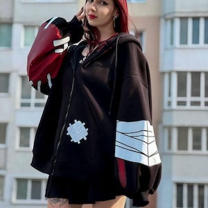 Op de afbeelding: Zwarte hoodie met rits en rode en witte accenten op de mouwen. De hoodie heeft een witte patch op de voorkant en een zwarte rok. De mouwen hebben een rood-wit ontwerp. De persoon draagt gestreepte kniekousen.