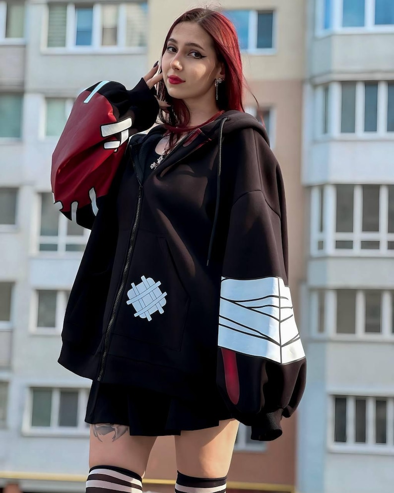 Op de afbeelding: Zwarte hoodie met rits en rode en witte accenten op de mouwen. De hoodie heeft een witte patch op de voorkant en een zwarte rok. De mouwen hebben een rood-wit ontwerp. De persoon draagt gestreepte kniekousen.