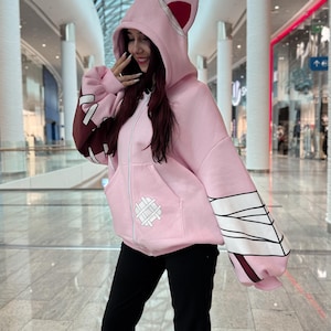 May include: A light pink zip-up hoodie with a hood featuring cat ears and a white zipper. The sleeves have a brown and white design. The hoodie has a front pocket with a white stitched design. The person is wearing black pants.
