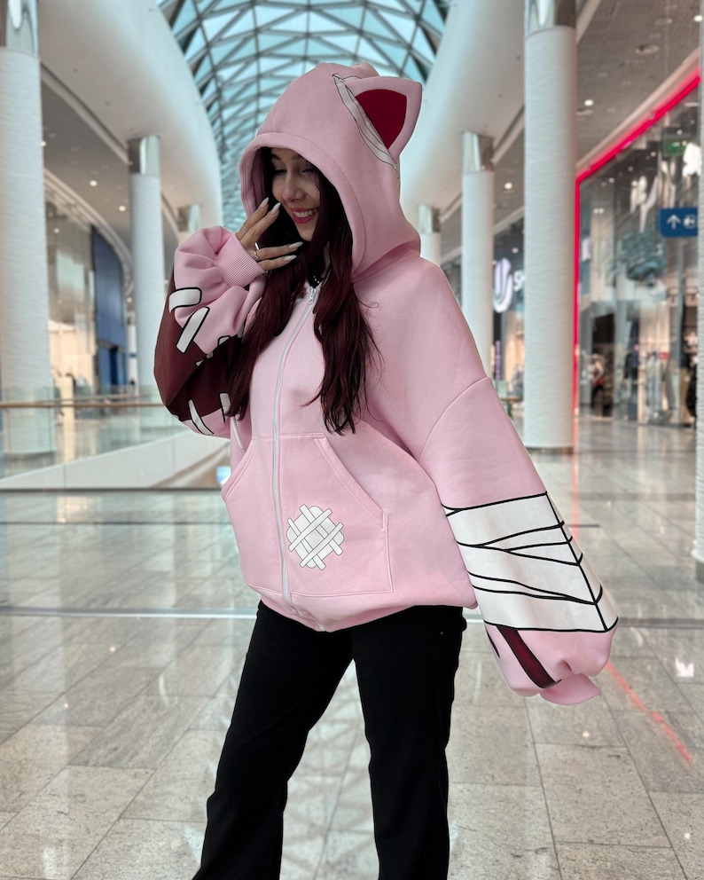 May include: A light pink zip-up hoodie with a hood featuring cat ears and a white zipper. The sleeves have a brown and white design. The hoodie has a front pocket with a white stitched design. The person is wearing black pants.