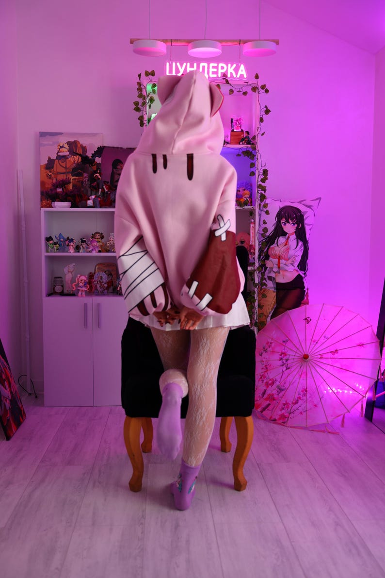 May include: A person wearing a pink hooded sweatshirt with bear ears, brown accents, and white bandages on the sleeves. The person is wearing white lace tights and purple boots. The word "ЦУНДЕРКА" is written in pink neon.