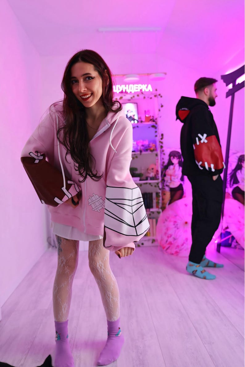 May include: A light pink zip-up hoodie with brown and white accents on the sleeves. The hoodie features a white graphic design on the sleeves and a white zipper. The person is wearing a white dress, white lace stockings, and purple socks.