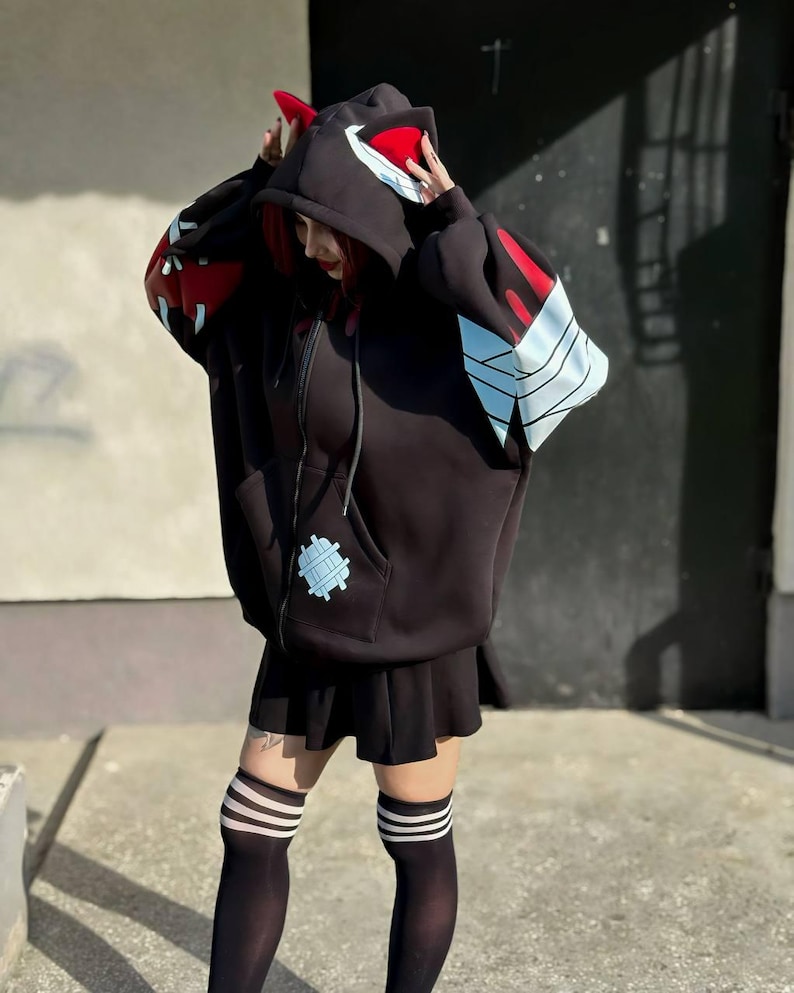 Op de afbeelding: Zwarte hoodie met ritssluiting en rode en witte accenten op de capuchon en mouwen. De hoodie heeft een uniek ontwerp met een patch op de voorzak. De outfit omvat een zwarte rok en kniehoge sokken met witte strepen.