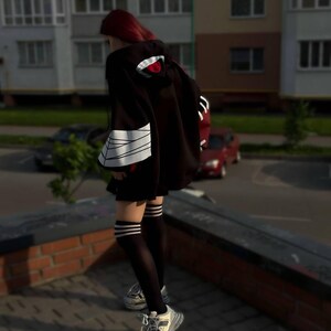 Op de afbeelding: Een persoon draagt een zwarte capuchon met een rood en wit ontwerp op de capuchon en mouwen. De persoon draagt ook een zwarte rok, kniehoge sokken met witte strepen en grijs-witte sneakers.