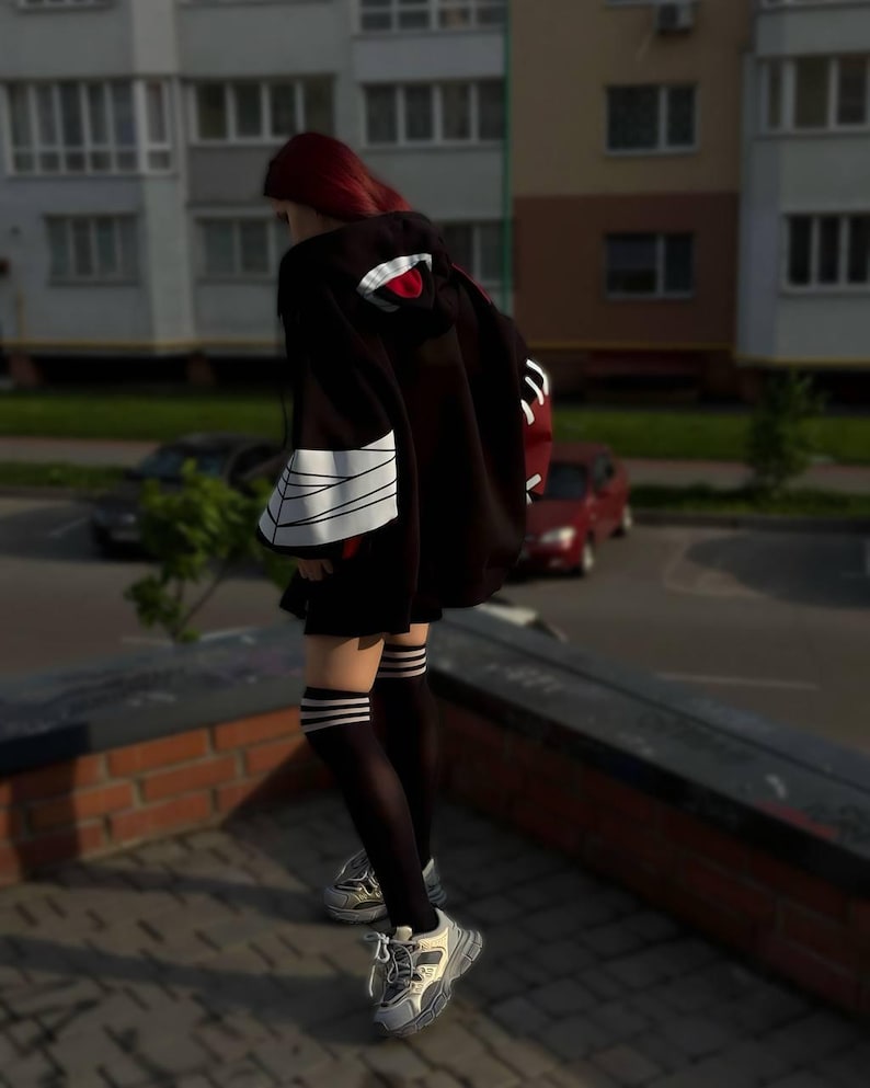 Op de afbeelding: Een persoon draagt een zwarte capuchon met een rood en wit ontwerp op de capuchon en mouwen. De persoon draagt ook een zwarte rok, kniehoge sokken met witte strepen en grijs-witte sneakers.