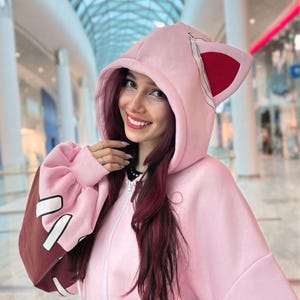 May include: A light pink zip-up hoodie with cat ears on the hood. The sleeves are a darker pink with brown accents and white shapes. The hoodie has a full zipper and a relaxed fit, perfect for casual wear.
