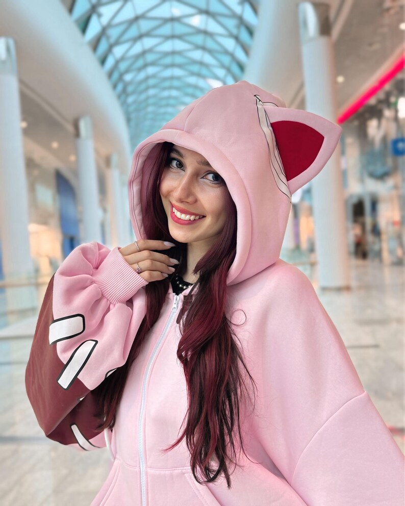 May include: A light pink zip-up hoodie with cat ears on the hood. The sleeves are a darker pink with brown accents and white shapes. The hoodie has a full zipper and a relaxed fit, perfect for casual wear.