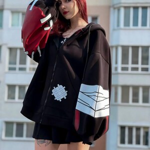 Op de afbeelding: Zwarte hoodie met ritssluiting en rode en witte accenten op de mouwen. De hoodie heeft een uniek ontwerp met een patch op de voorkant en een opvallende grafische print op de mouwen. Het model draagt een zwarte rok en gestreepte sokken.