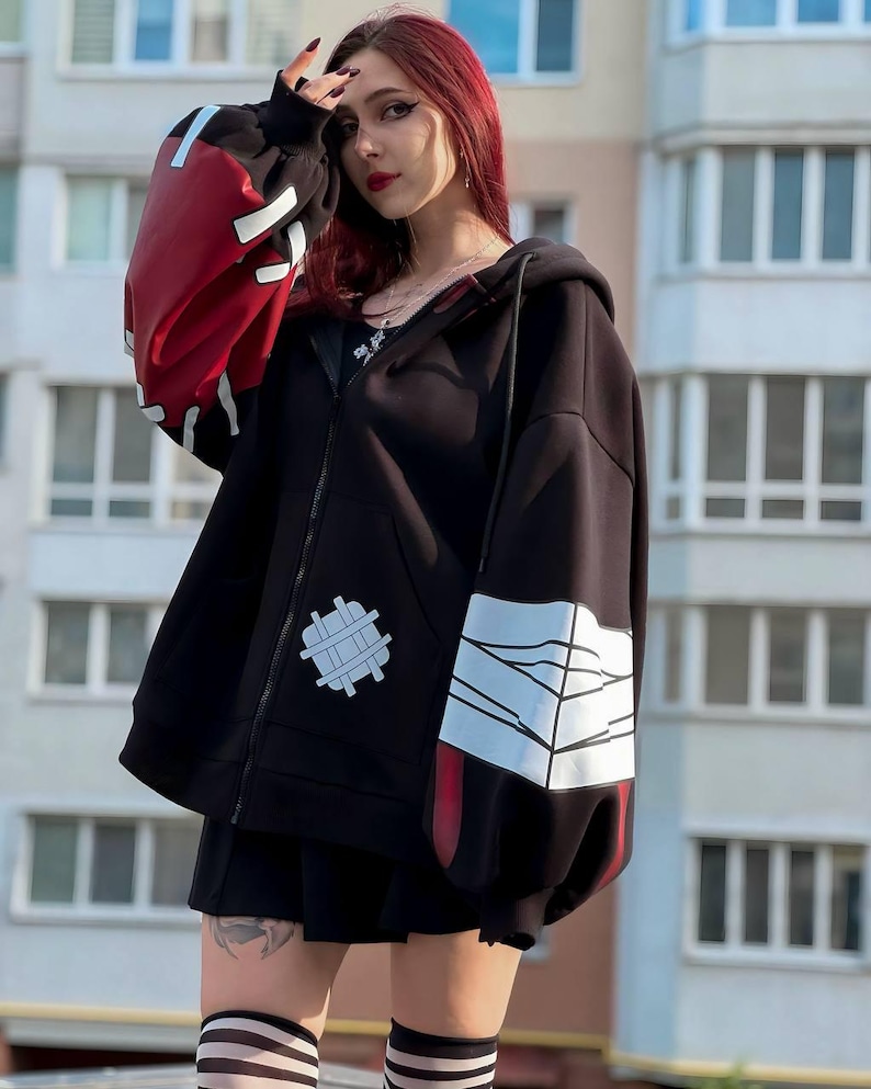 Op de afbeelding: Zwarte hoodie met ritssluiting en rode en witte accenten op de mouwen. De hoodie heeft een uniek ontwerp met een patch op de voorkant en een opvallende grafische print op de mouwen. Het model draagt een zwarte rok en gestreepte sokken.