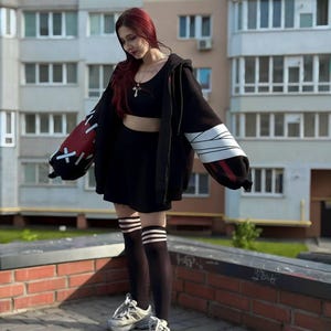 Op de afbeelding: Een zwarte outfit bestaande uit een crop top, rok en oversized hoodie met rode en witte accenten op de mouwen. De outfit wordt gecombineerd met zwarte kniehoge sokken met witte strepen en witte sneakers.