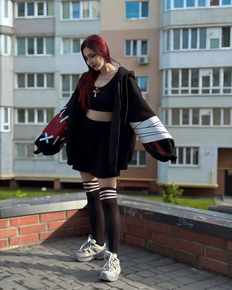 Op de afbeelding: Een zwarte outfit bestaande uit een crop top, rok en oversized hoodie met rode en witte accenten op de mouwen. De outfit wordt gecombineerd met zwarte kniehoge sokken met witte strepen en witte sneakers.