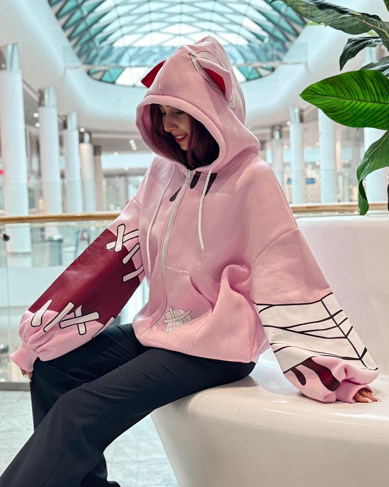 May include: Pink zip-up hoodie with a hood featuring red accents and ears. The sleeves have burgundy and white designs. The hoodie has a front pocket and white drawstrings. The person is wearing black pants.